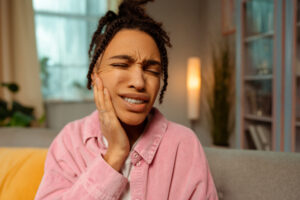 Woman in pink shirt experiencing tooth pain, holding her cheek with eyes closed.