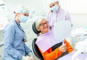 Senior woman smiling in dental chair with two hygienists during implant consultation at Hawthorn Woods practice.